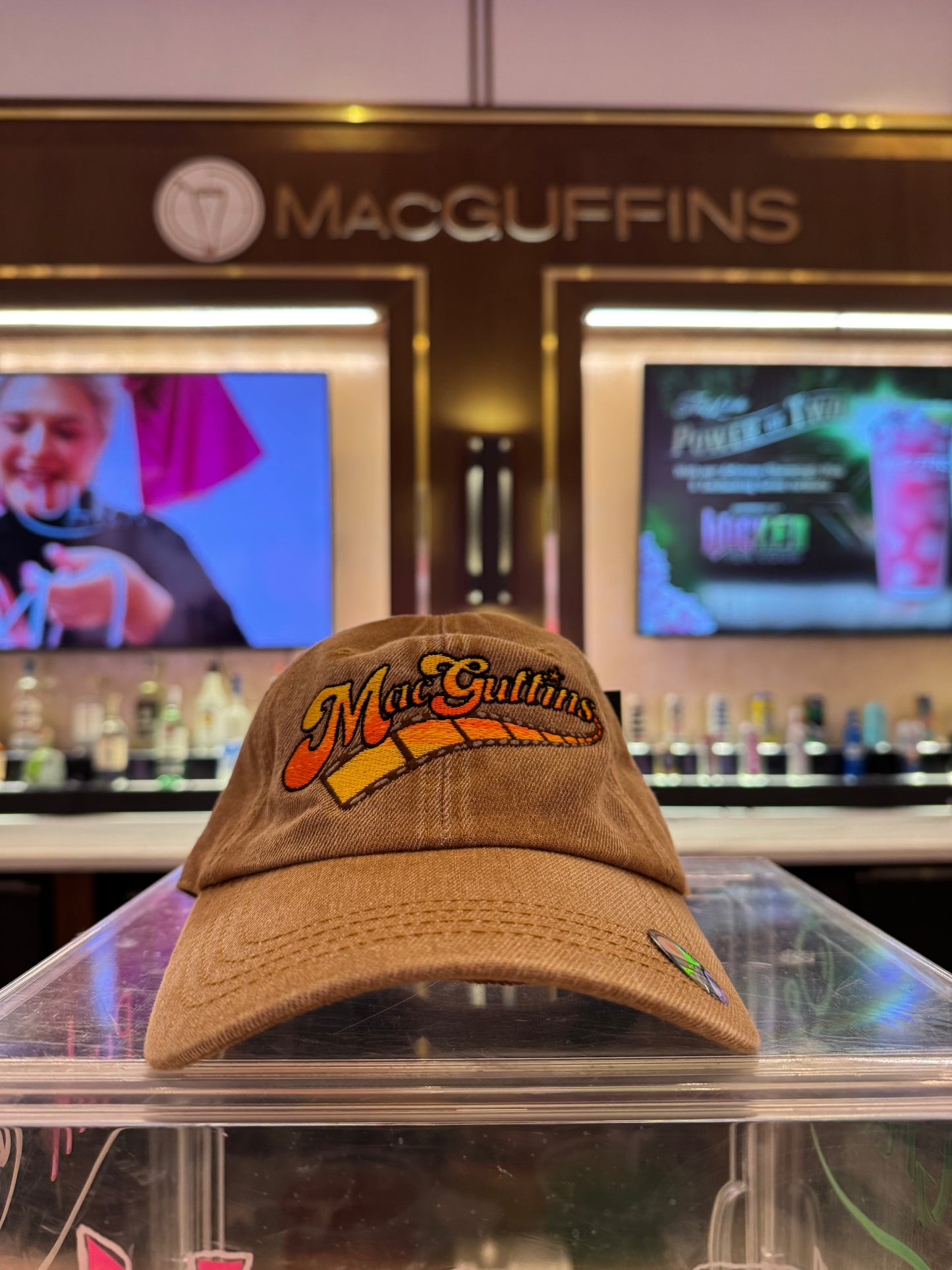 Brown cap with 'MacGuffins' logo on a glass surface with a bar background.